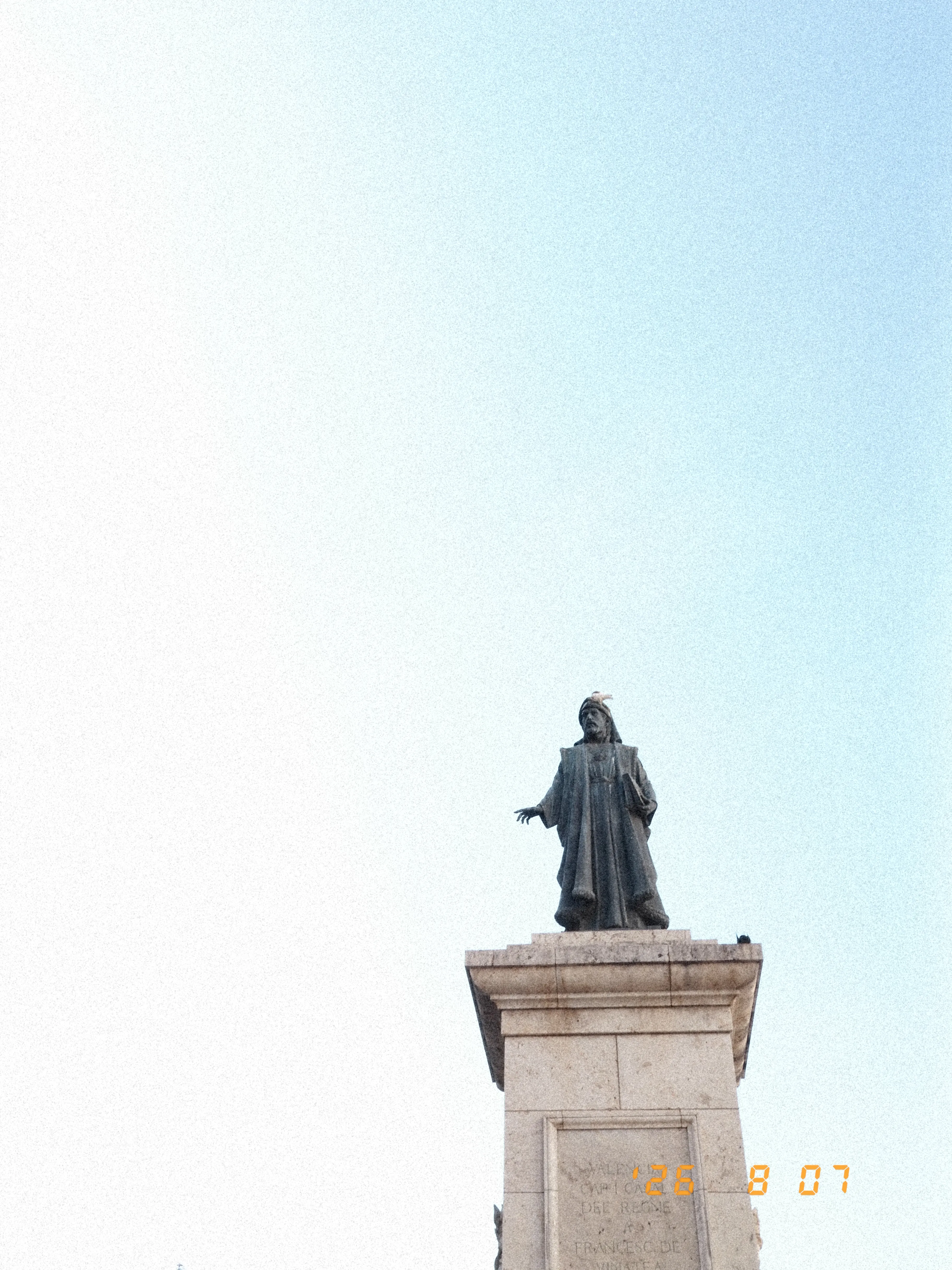 A grey statue of a robed man with a white bird on his head, surrounded by blue sky