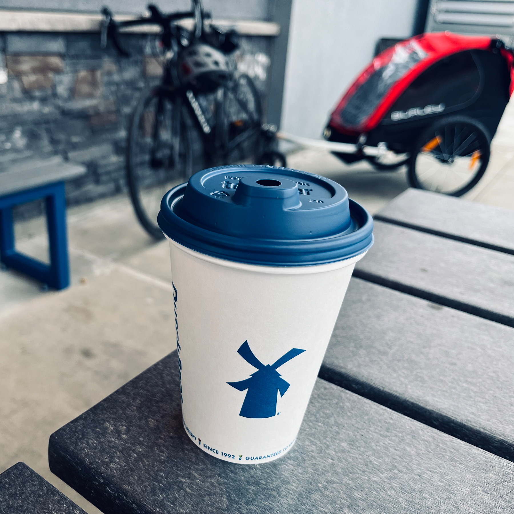 A disposable coffee cup with a blue lid and a windmill logo on the side rests on an outdoor table, with a blurred background featuring a bench, a bicycle, and a bike trailer.