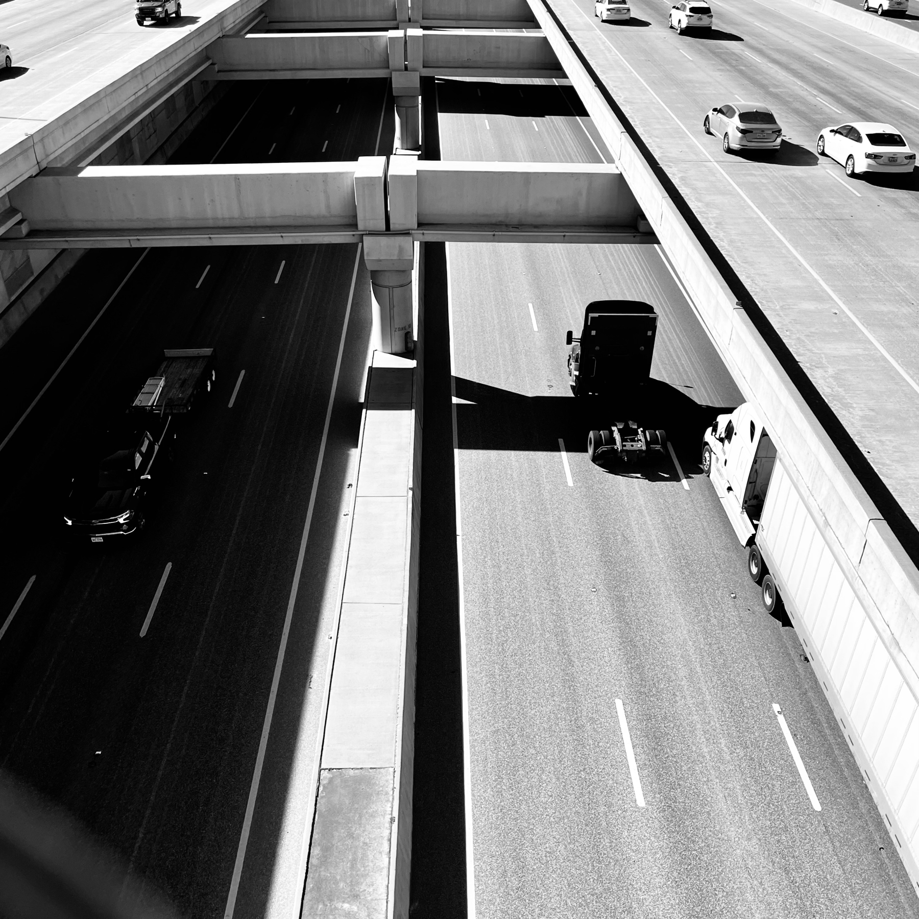 Black and white image of a multi-level highway system with cars and trucks driving on the sunlit roads, casting sharp shadows from the overhead structure